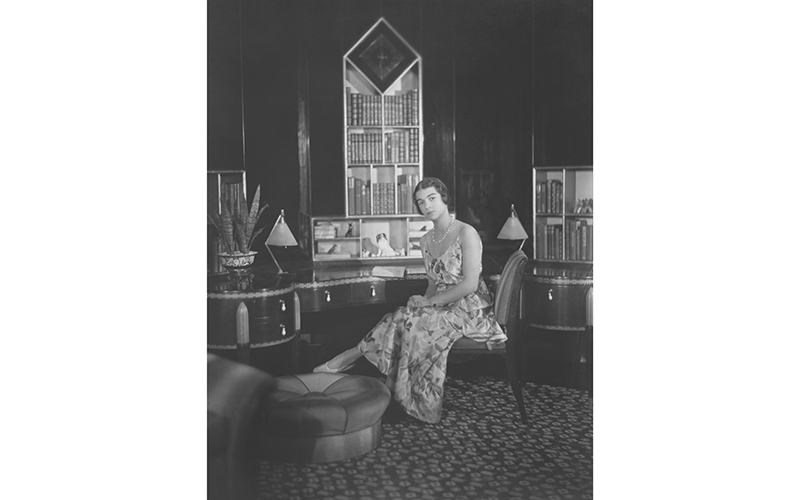 Elaine Wormser, dressed in a floral cocktail dress, sitting at her desk in her bedroom, looking toward us.