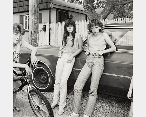 Two cool-looking women lean on a car. A boy rests on a bike to their side.