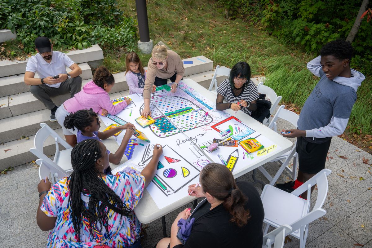 An outdoor view, looking down from above, of a large square table surrounded by eight people participating in art-making activities.