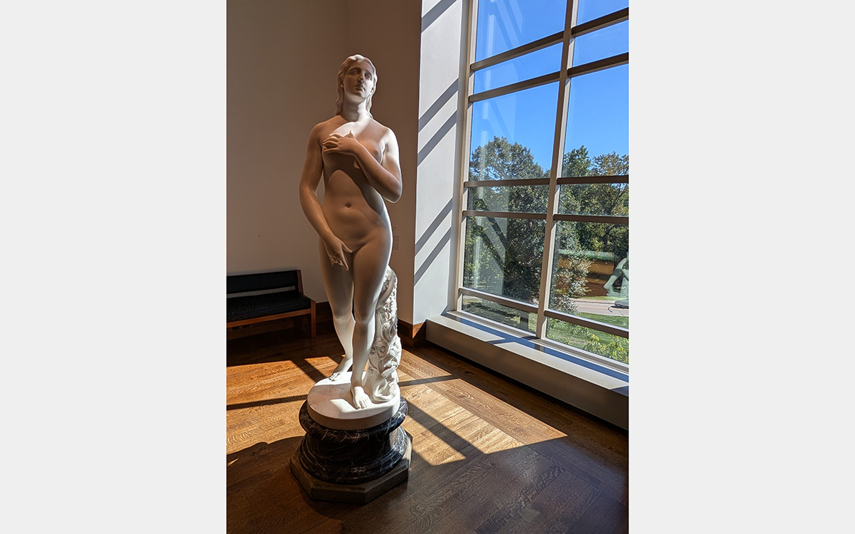 White marble sculpture of a nude woman by a floor-to-ceiling window overlooking a park-like setting and blue sky.
