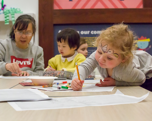 A young person leans on a table in the REC, intently focused on their drawing