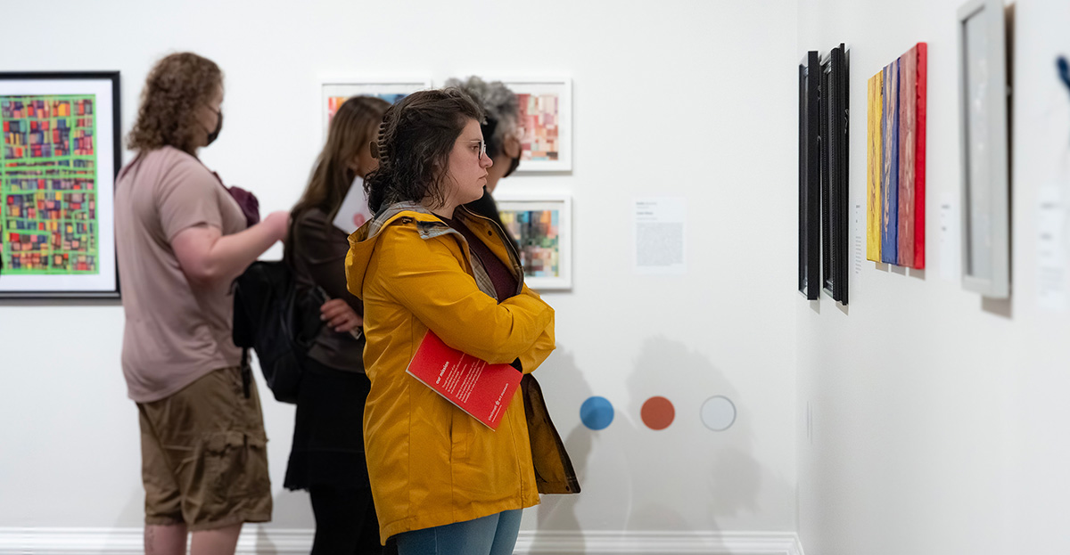A white woman in a yellow jacket looks at framed art on a wall