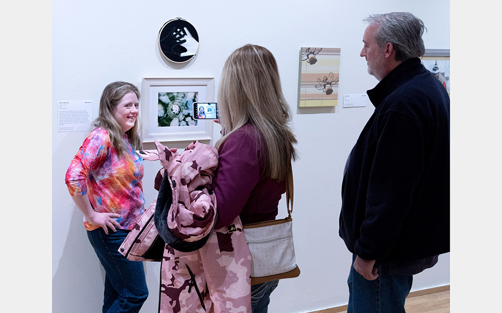 A white woman smiles and gets her picture taken beside a small green and white framed artwork.