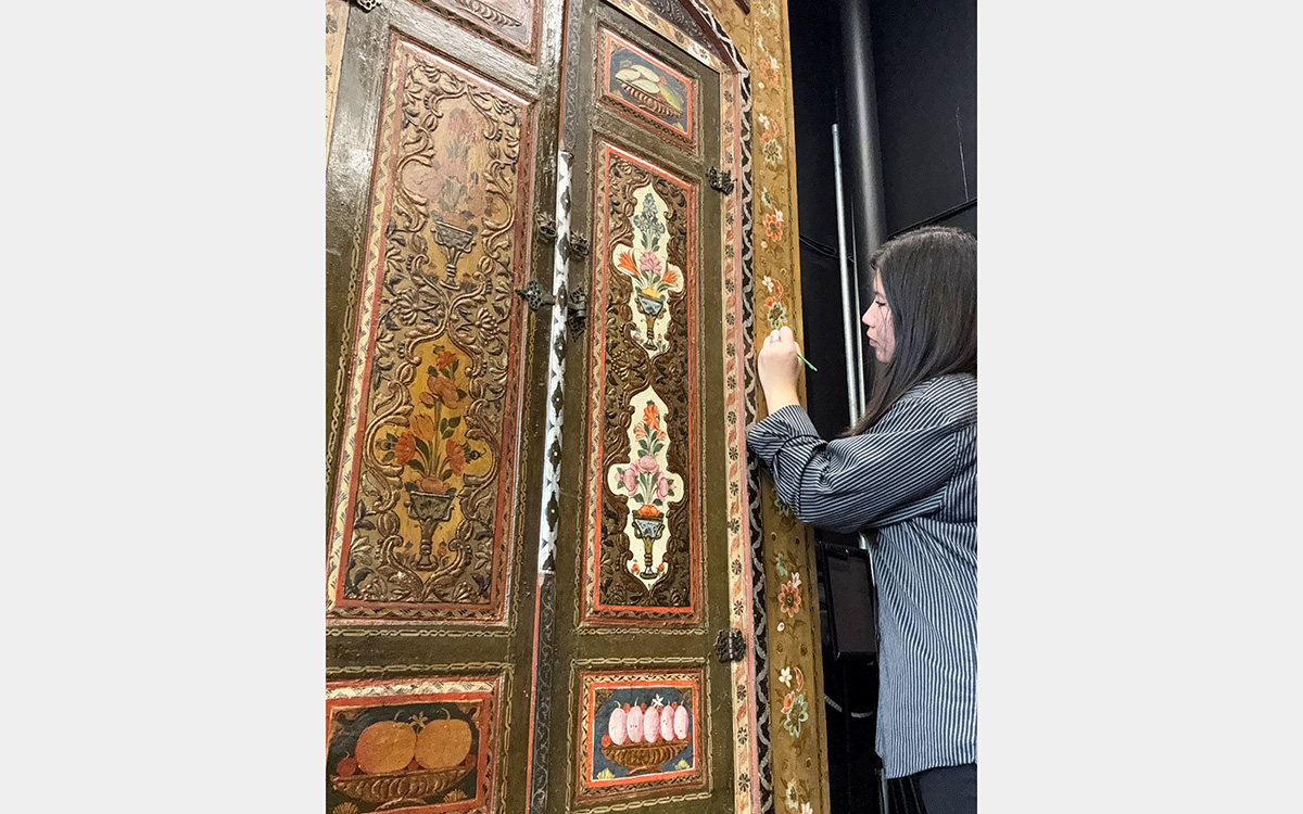 A woman uses a small tool to remove old varnish from a decorative wooden wall panel.