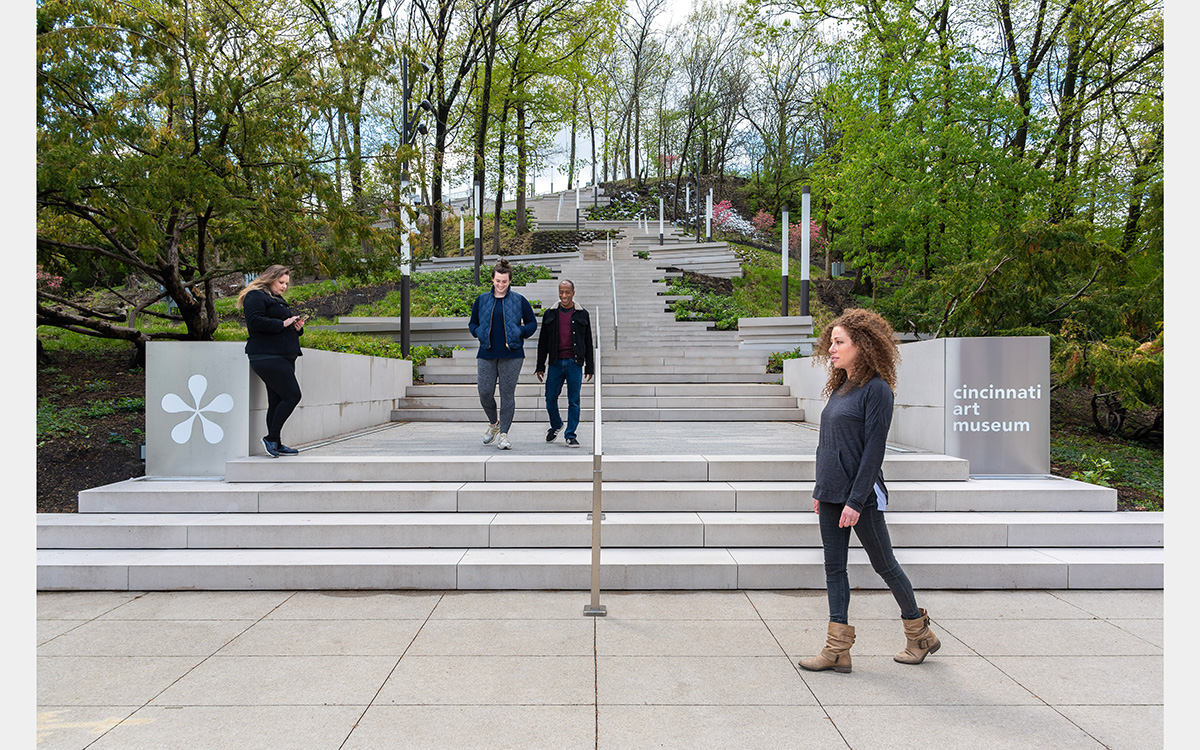 People walking at the bottom of limestone-colored stairs that go up a tree-filled hillside.