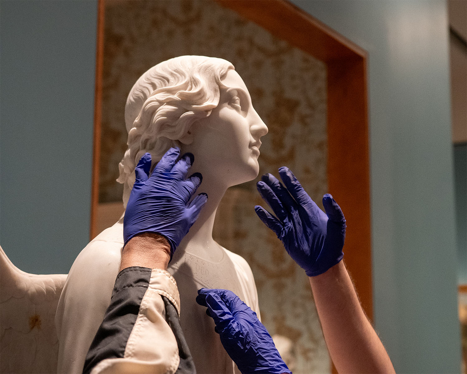 Gloved hands reach up to touch the face of an angel sculpture