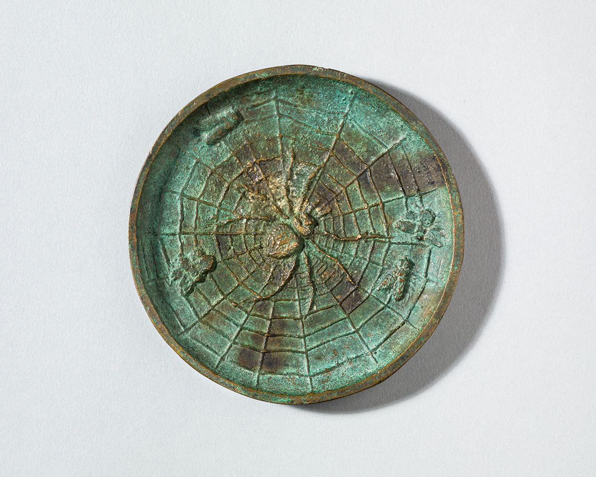 Circular bronze object with relief of a spider, spider web, and insects.