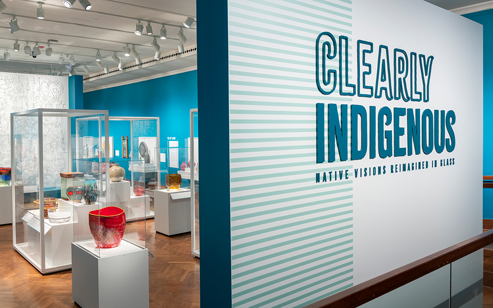 A large wall with the text "Clearly Indigenous: Native Visions Reimagined in Glass," with cases filled with colorful artworks in the background.