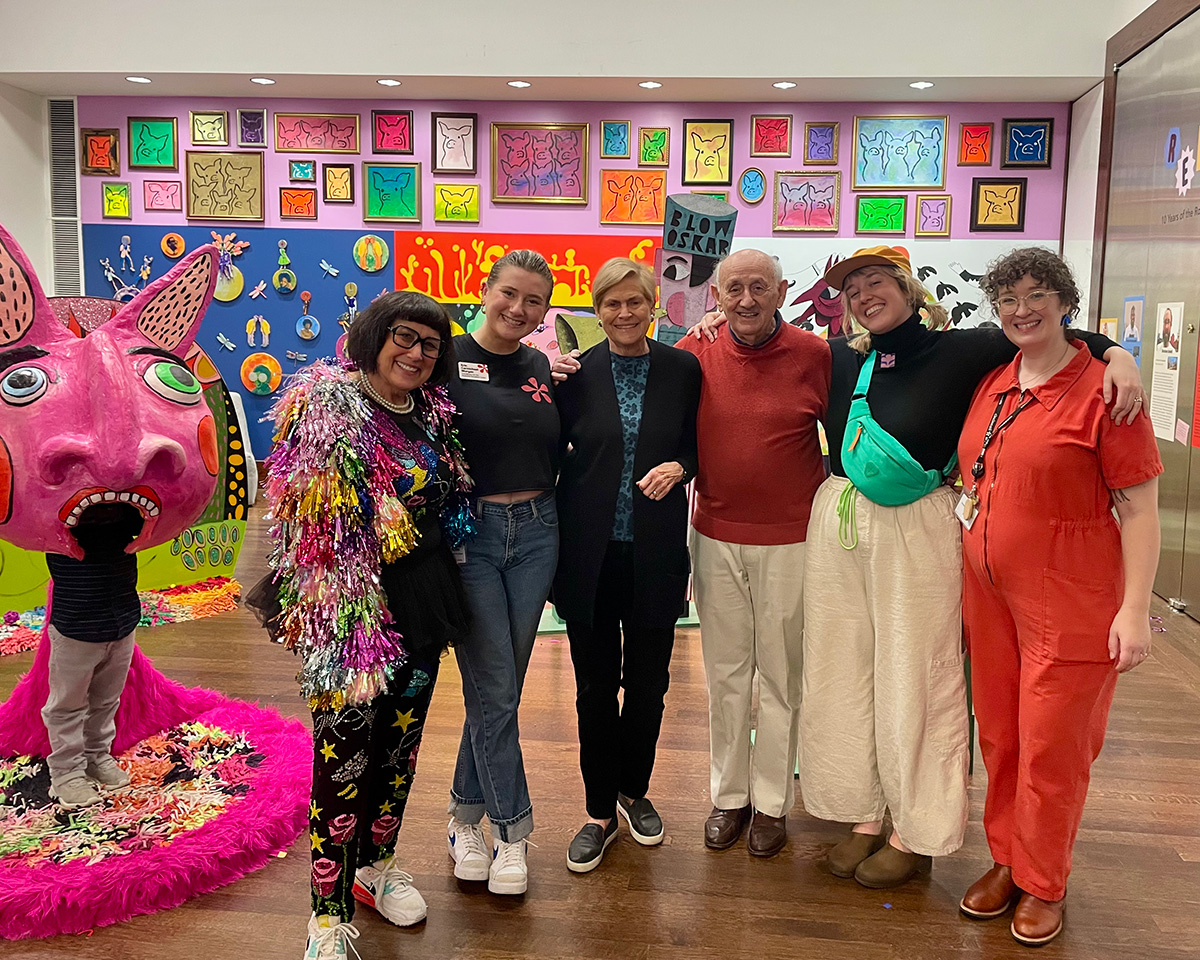 A group of six colorfully dressed people stand in a room in a room with a brightly colored wall in the background. To the left of the group, a child stands wearing a large pink mask with pointed ears.