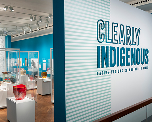 A large wall with the text "Clearly Indigenous: Native Visions Reimagined in Glass," with cases filled with colorful artworks in the background.