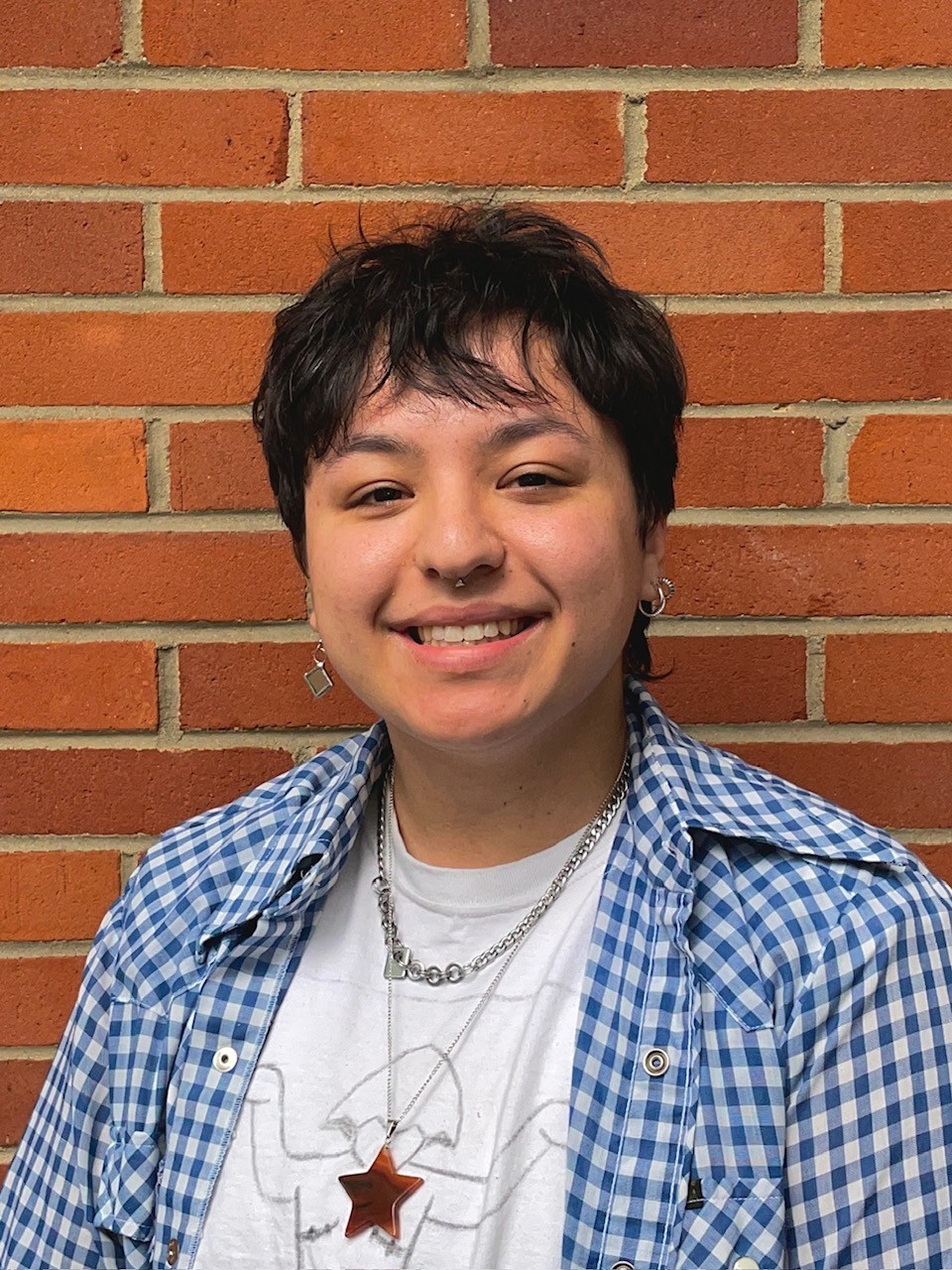 Photo of a person smiling at the camera, standing against a brick wall, wearing a blue button down shirt.