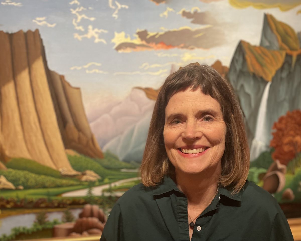Smiling woman stands in front of a landscape painting.