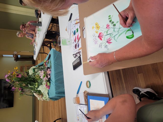 A woman paints flowers on paper at a U-shaped table with a flower arrangement in center of room.