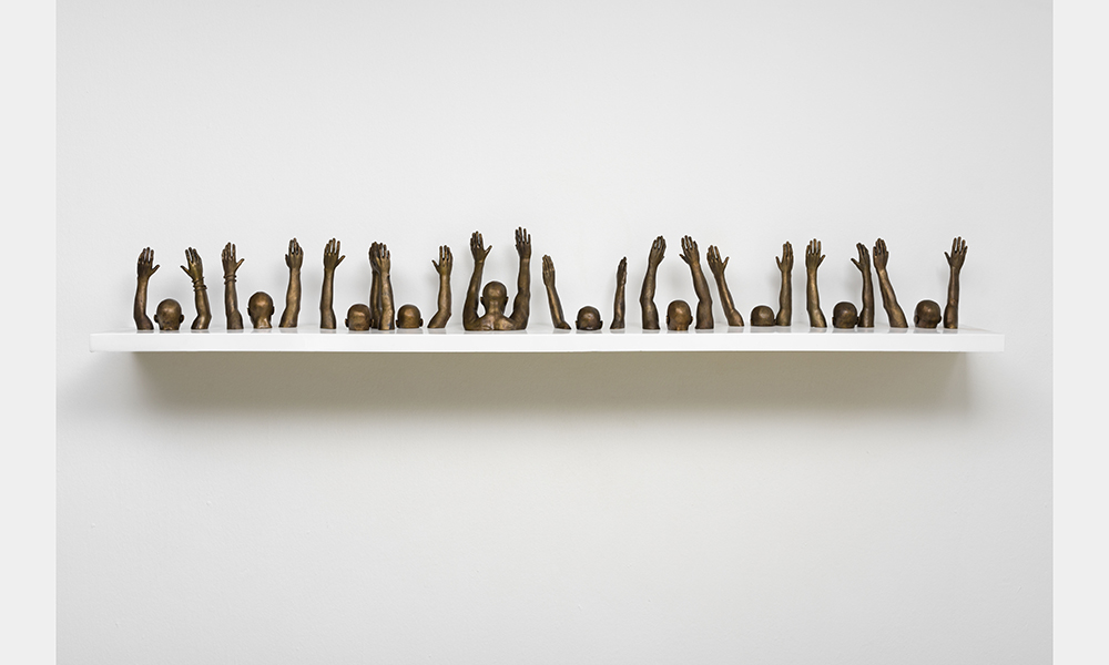 Hank Willis Thomas' Raise Up, a bronze statuette of a line of people with their hands raised above their heads facing away from the viewer. The statues are only depicted from the shoulders up, seemingly sinking into the shelf on which they rest