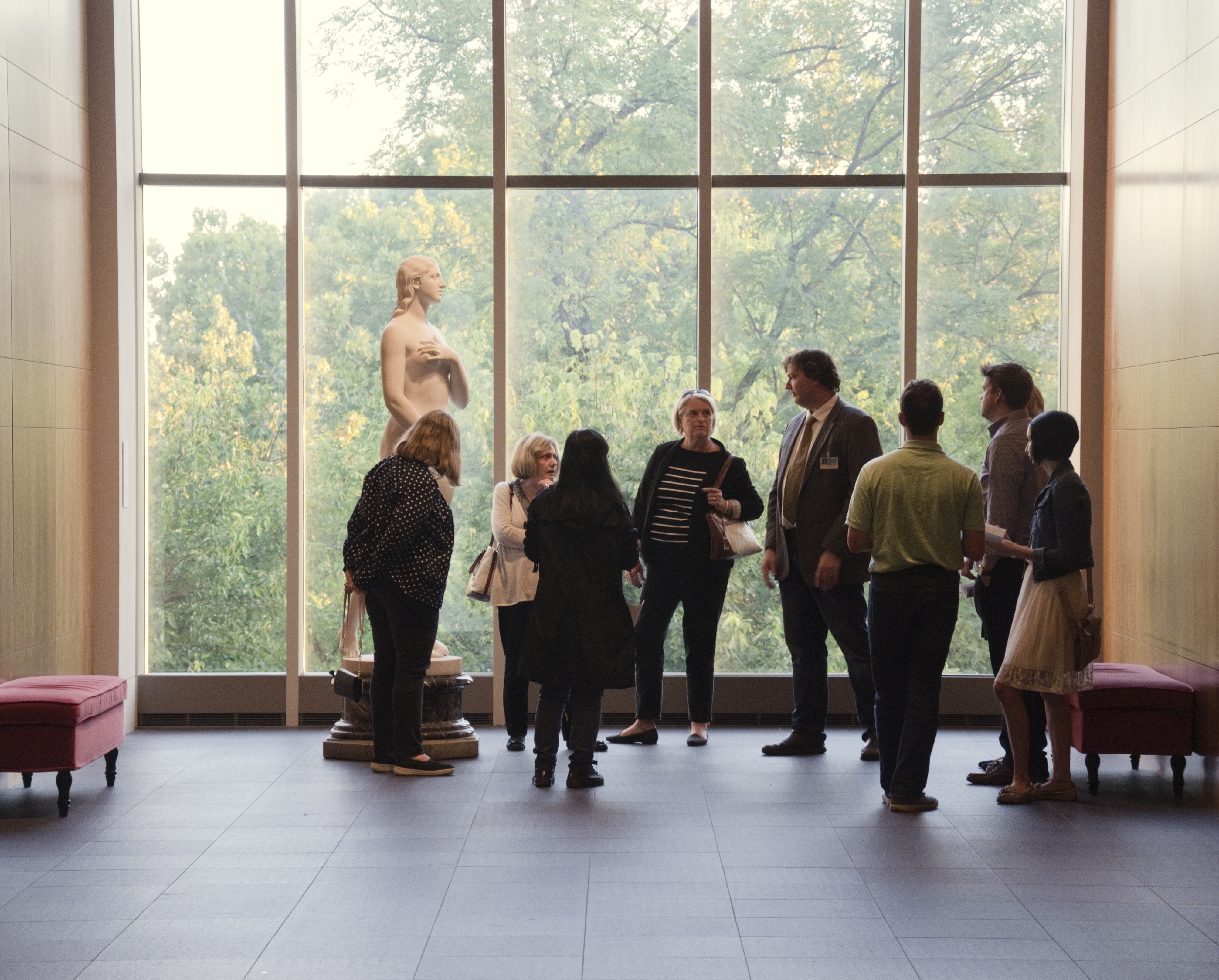 A group of people stands near a marble statue of the woman with a large window in the background with a view of green trees.