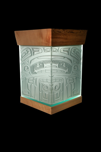 The sculpture consists of a rectangular box of tinted green glass which has a warm brown cedar base and lid. The lid is slightly wider and flared compared to the thinner, more squared-off base. The object is angled so that a corner of the box faces the viewer. This corner bisects a symmetrical design that has been sandblasted into the surface of the glass. The rounded geometric shapes that comprise the design may represent the face and body of an animal-like figure, perhaps that of the mouse in the work’s title.