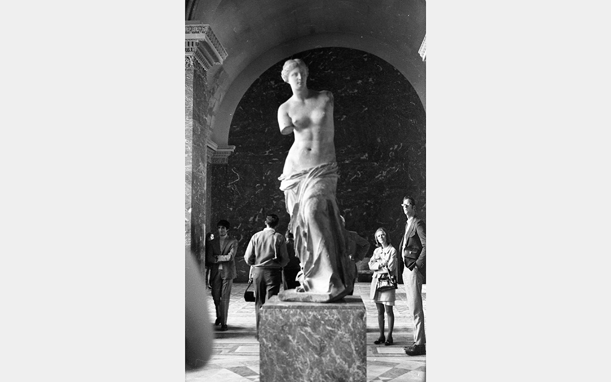 A white marble statue representing a woman; both arms are missing; lower half is covered by drapery; upper half is nude; hair is pulled back.