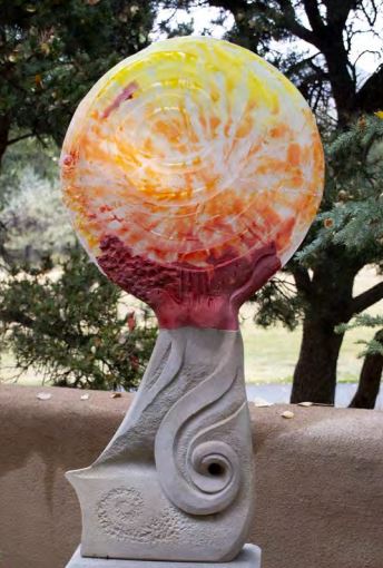 A large round glass sculpture with yellows, oranges and reds on a stone base
