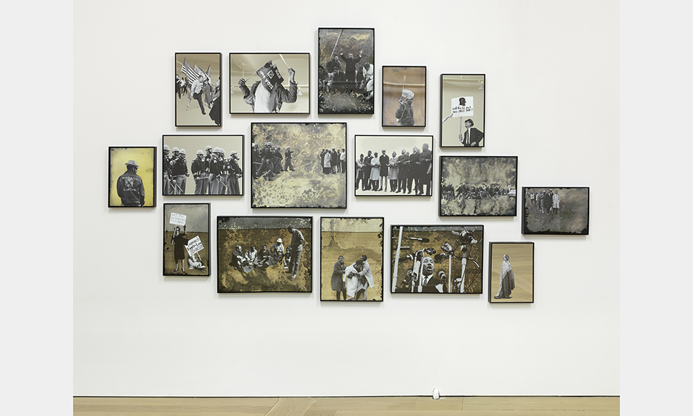 Hank Willis Thomas' Ain't Gonna Let Nobody Turn Us Around, a series of framed, rectangular screen prints arranged in a collage on the wall featuring images of Civil Rights Era civil rights activists, counter protestors, and riot police in front of yellow and beige backdrops