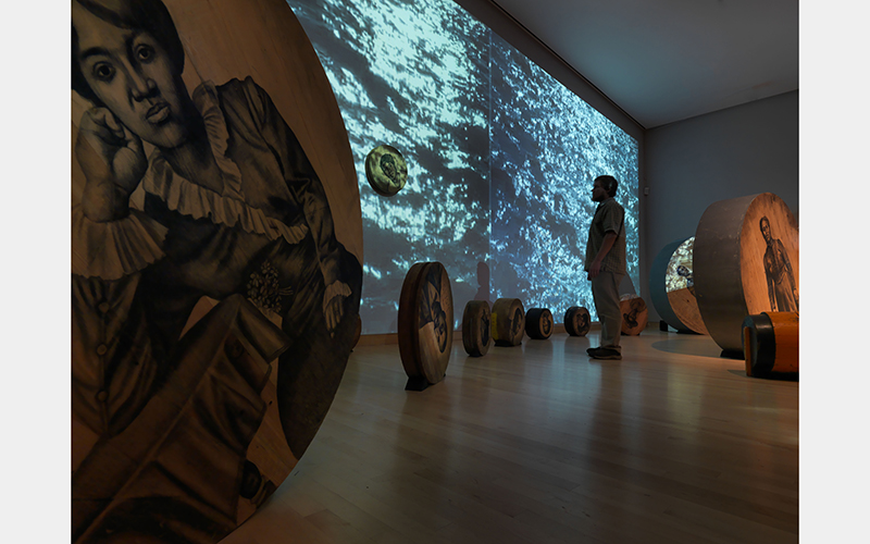 A person stands in a room filled with wooden circles and a projection of moving water. The wooden circles have monochromatic portraits of people on them.