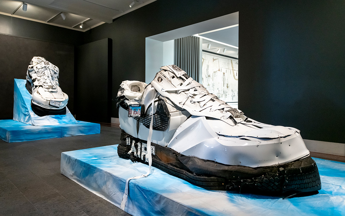 Large-scale metal sculptures representing a pair of sneakers displayed inside a gallery space with visitors and a security guard sitting in a chair
