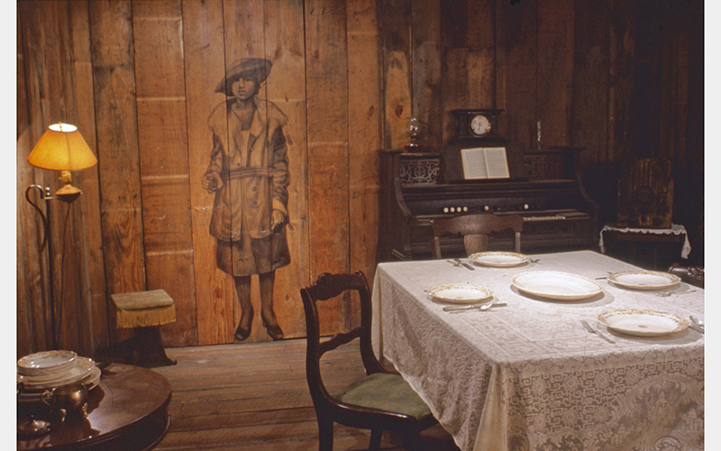 An illustration of a person wearing a hat and coat in a wood-paneled room with a piano, lamp, and dining table.