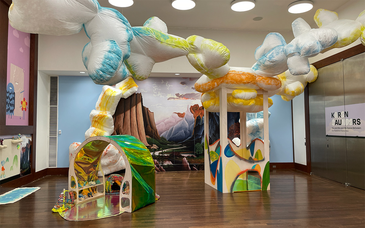 Fluffy blue and yellow clouds descend from the ceiling in front of a mural of a river between mountains