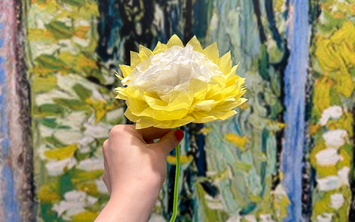 Human hand holds a bright yellow and white tissue paper flower against a colorful painted background.