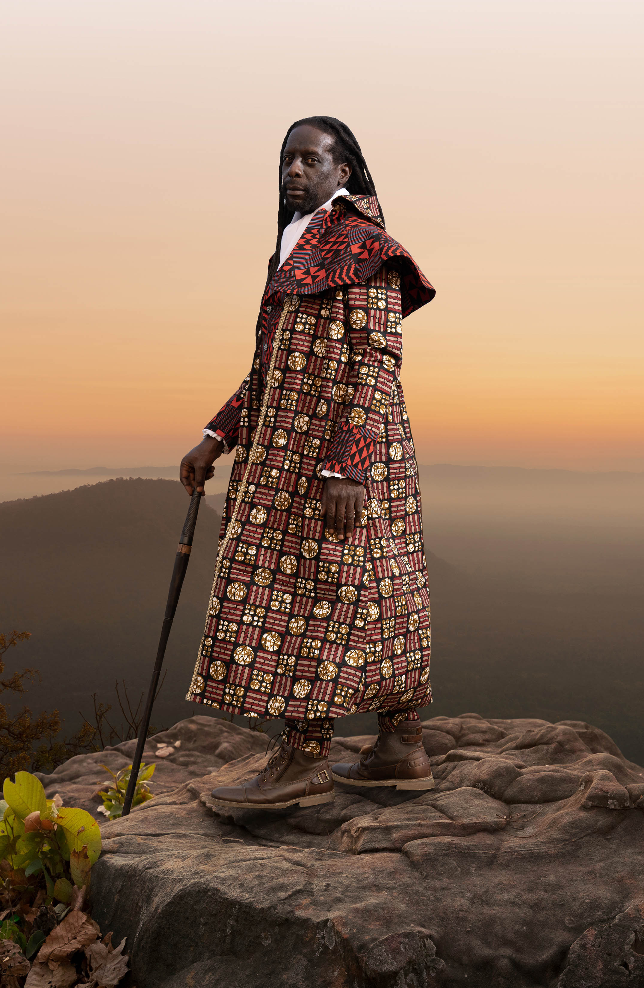 A Black man wears a red robe and stands on top of a mountain at sunset