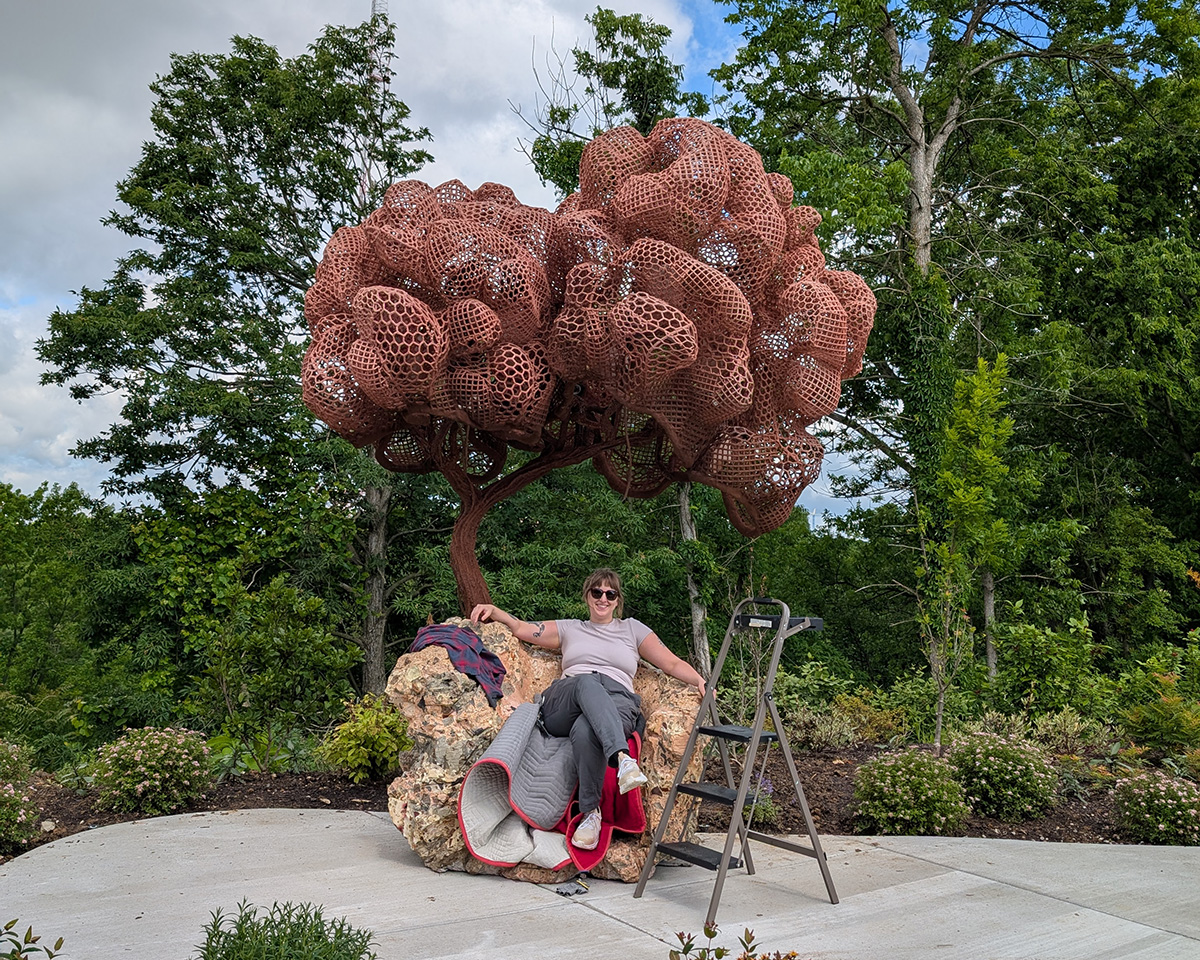 A woman sits under a brown tree-like on a red and gray blanket spread over a large rock-like form form. In the background is a green landscape. Next to the woman stands a stepladder. 