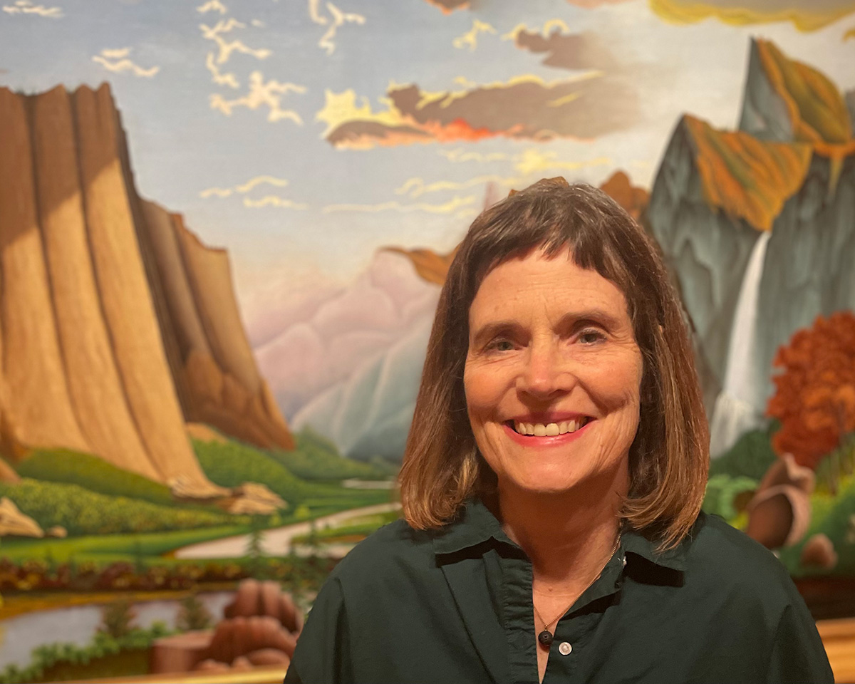 A woman in a green shirt with shoulder-length brown hair is pictured from her shoulders up in front of a colorful painting of a landscape.