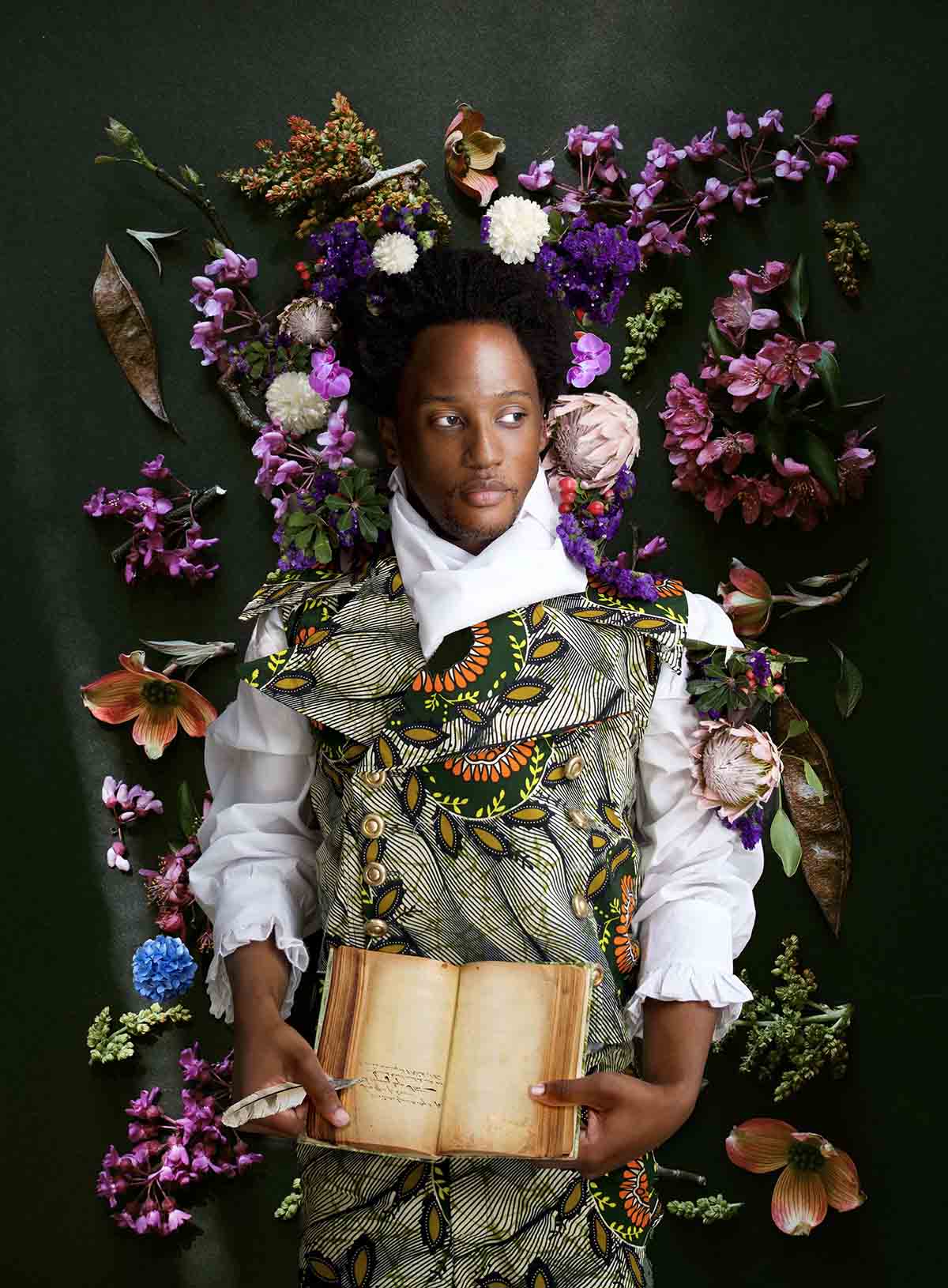 A Black man surrounded by flowers holds a book and a quill pen
