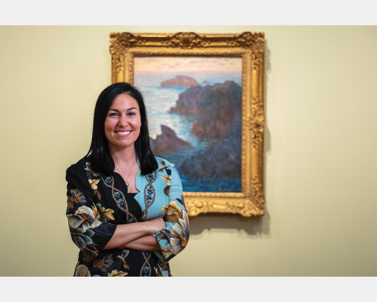 A smiling woman with shoulder-length black hair stands with in front of a painting in a gold frame depicting a seascape.
