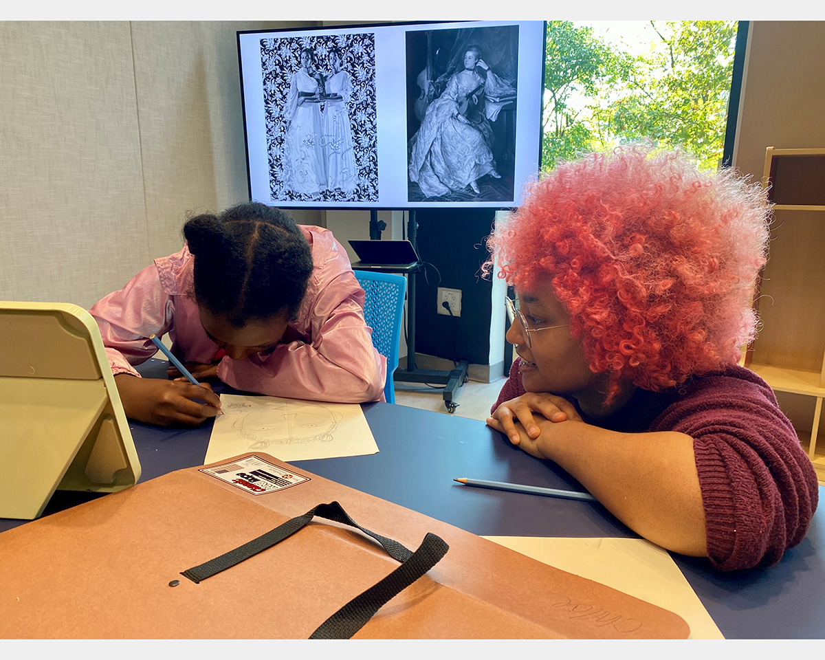 Two girls sit at a table while one draws. Black and white images of paintings are projected onto a screen in the background.