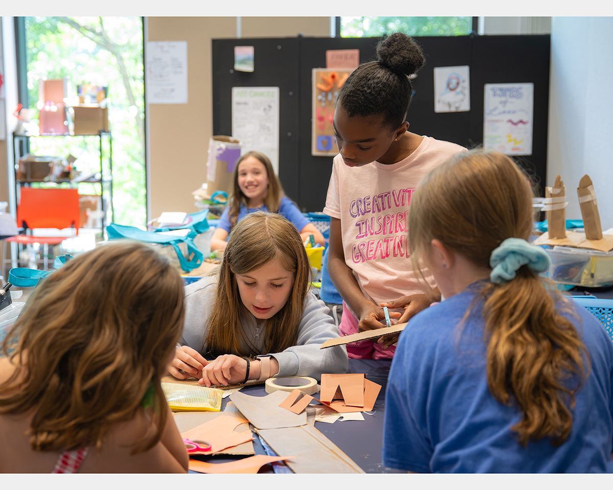 Four girls gather around a table while a fifth girl smiles in the background.