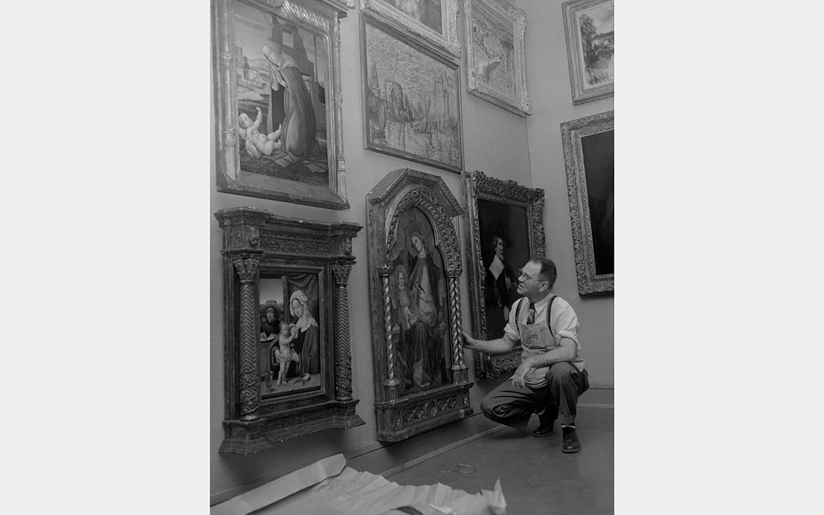 A black and white photo of a man crouching beside a framed painting in a gallery
