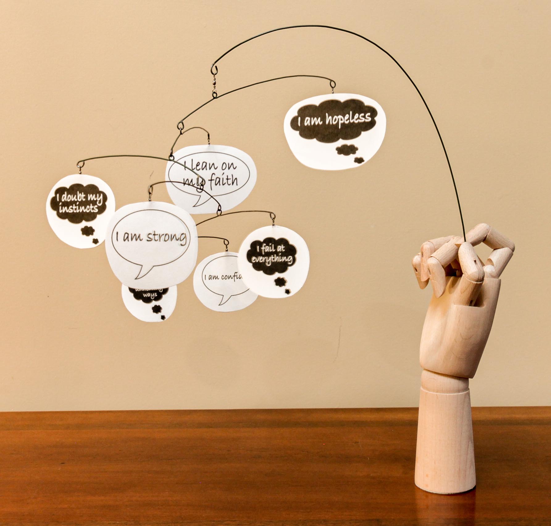 A wooden model hand holding a mobile with floating pieces with speech bubbles that say phrases such as "I am strong." "I am hopeless." "I am confidant." "I fail at everything." The positive affirmations are in white bubbles and the negative in black bubbles.