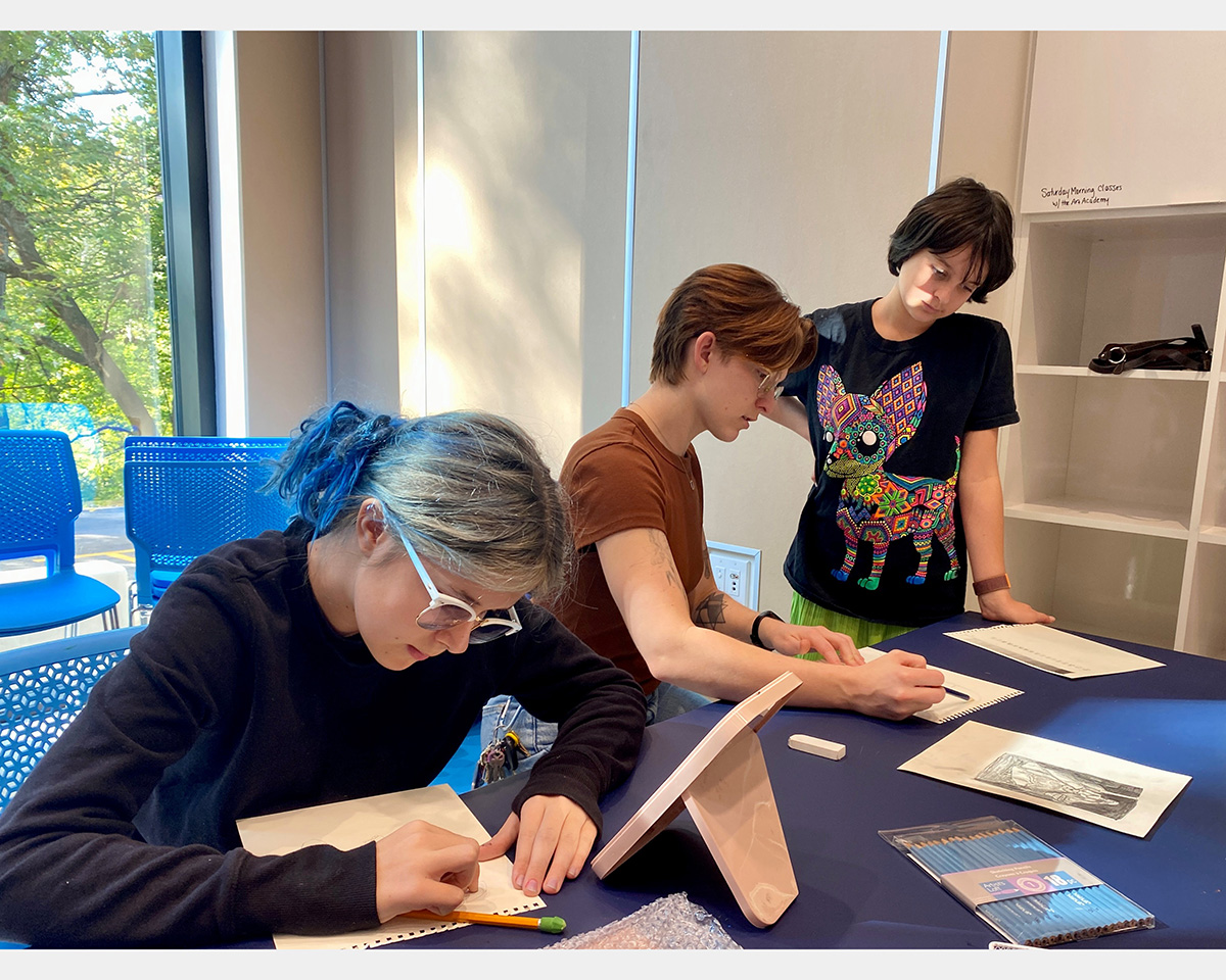 Two girls sit at a table making sketches while a third girl stands and watches.