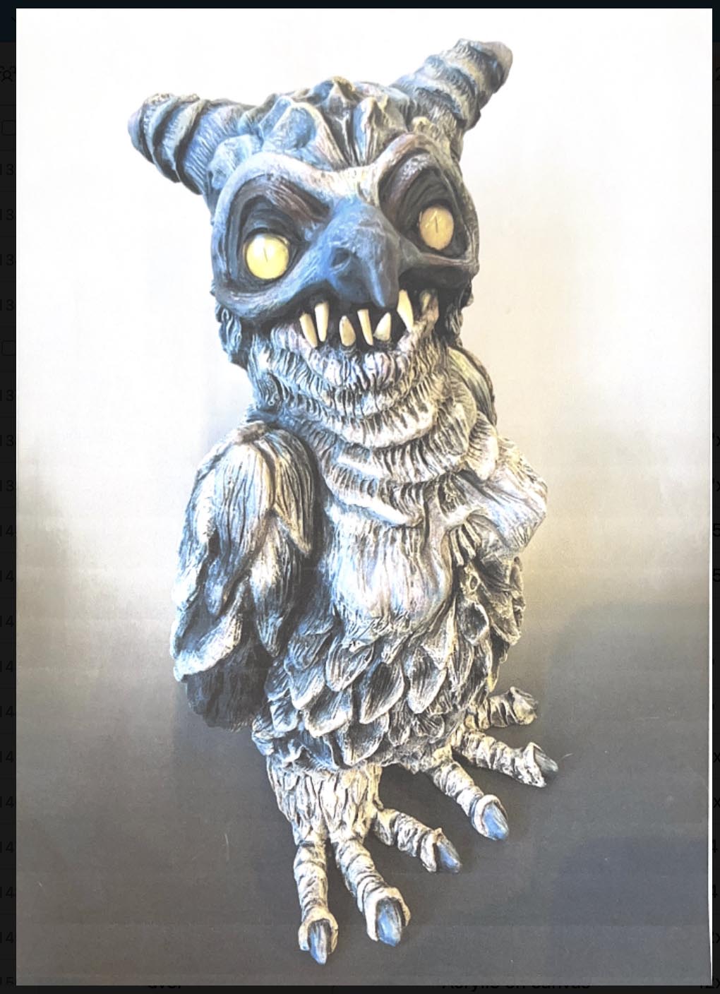 An owl-like creature with glowing yellow eyes and sharp dog-like teeth gazes menacingly at the viewer, the creature is a ceramic statue with intricate feather detailing and carefully sculpted appendages. The piece has a mystical, monster-like quality.