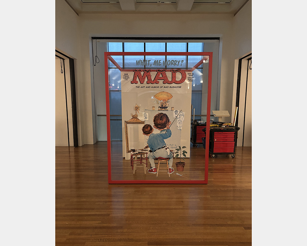 Large transparent sign in red frame with large text stating "MAD", displayed in space with tall ceilings, hardwood floors, and translucent window in background. 