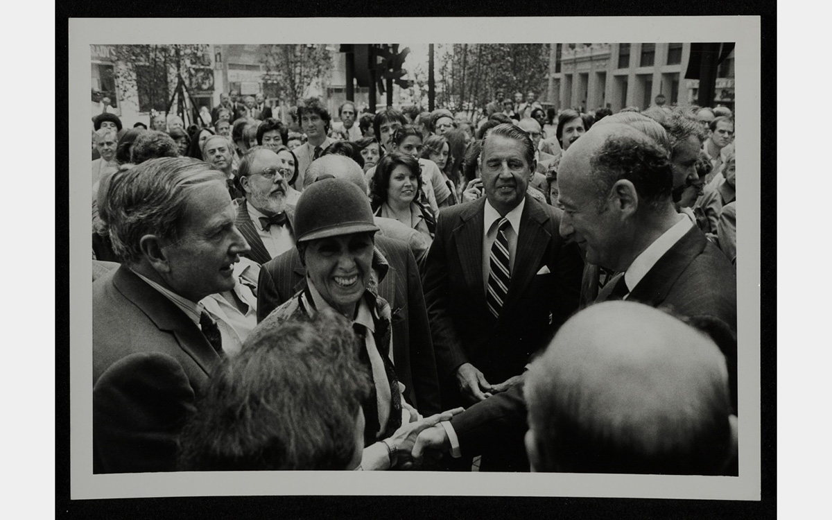 Black and white photo of a smiling woman shaking hands with a man surrounded by a crowd of people