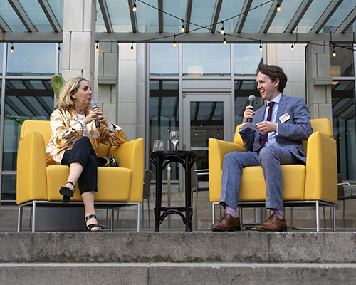 Two white people talk to each other while sitting in bright yellow chairs outdoors