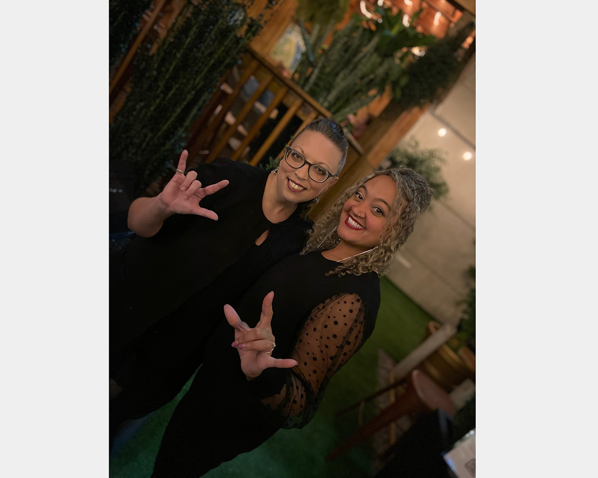 Two women, both dressed in black, stand against a green background giving hand signs. Camera angle is at a slant.