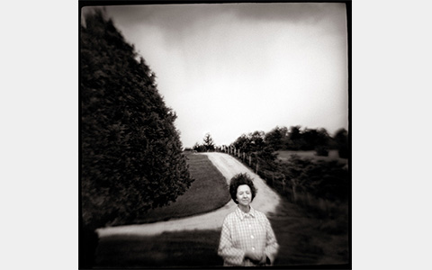 Nancy Rexroth's My Mother, black and white photograph of a woman standing in front of a dirt road curving around a large evergreen tree