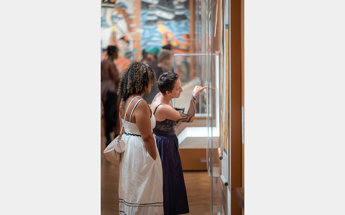 Two women looking at plexiglass wall case inside an exhibition. One points at something inside the case.