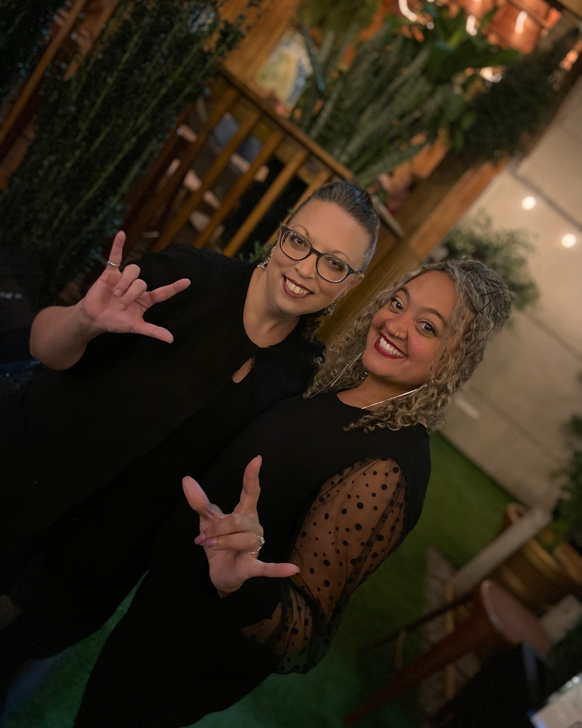 Two women, both dressed in black, stand against a green background giving hand signs. Camera angle is at a slant.