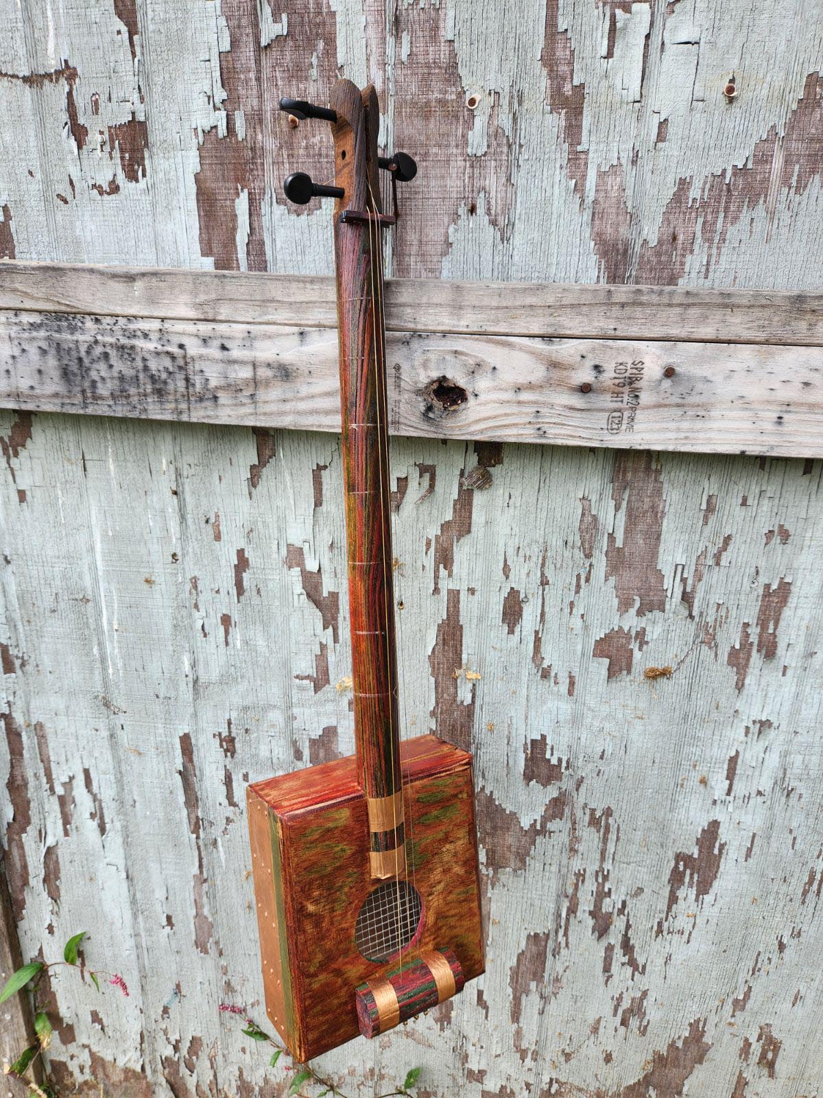 A three-string cigar box instrument with rich redwood with green accents gently blended in. The piece has a vintage folk feel to it.