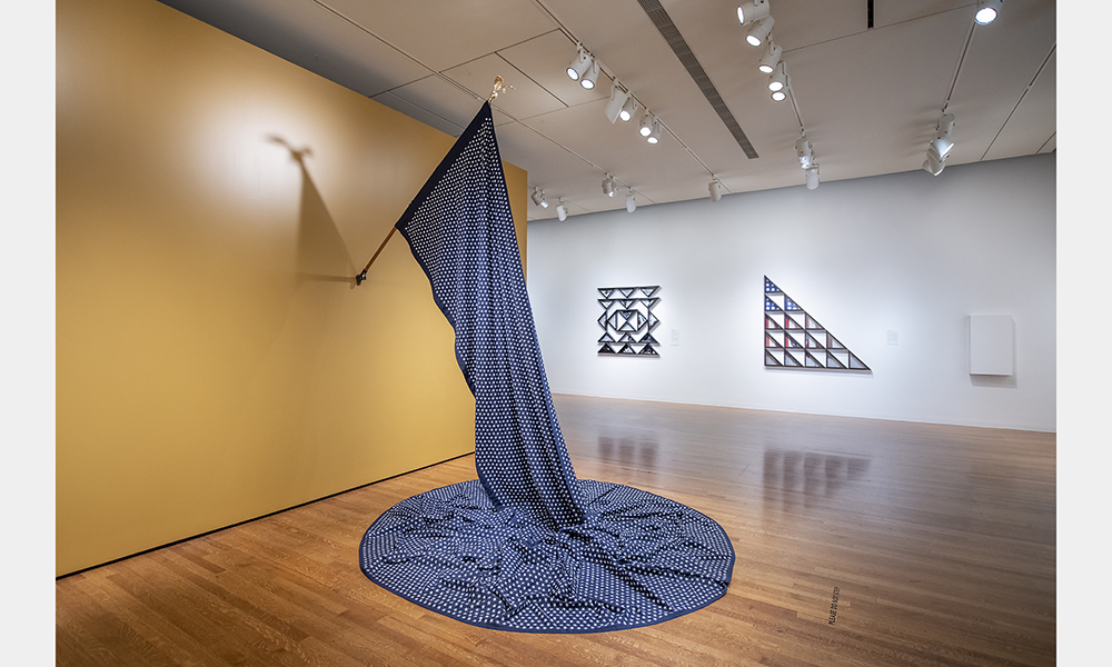 Hank Willis Thomas' 15,580, a large blue flag embroidered with white stars mounted on the wall. The flag droops down onto the floor into a circle like a large puddle