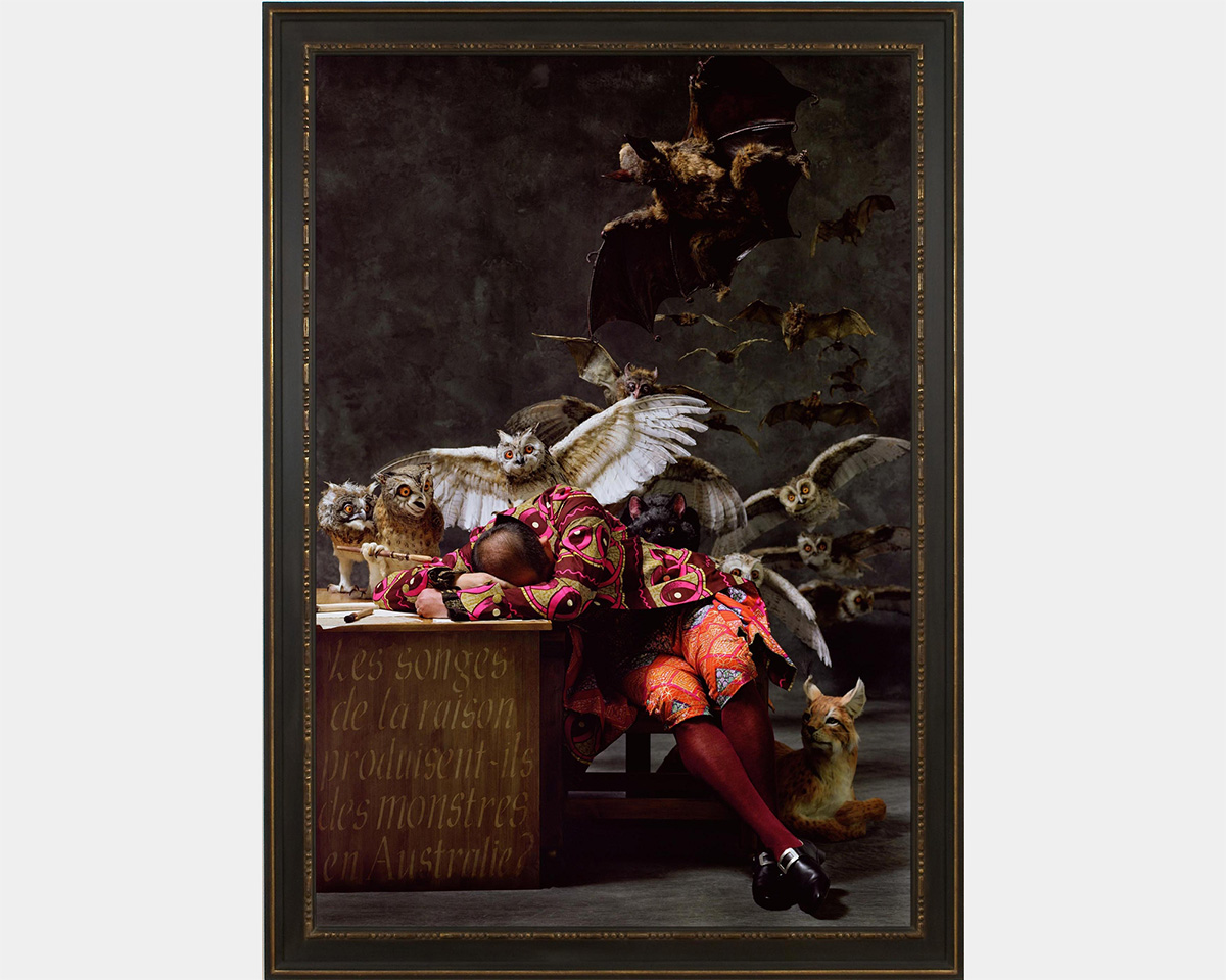 A man in a colorful outfit rests his head on a desk while owls and bats fly around him and a lynx lays by his side.