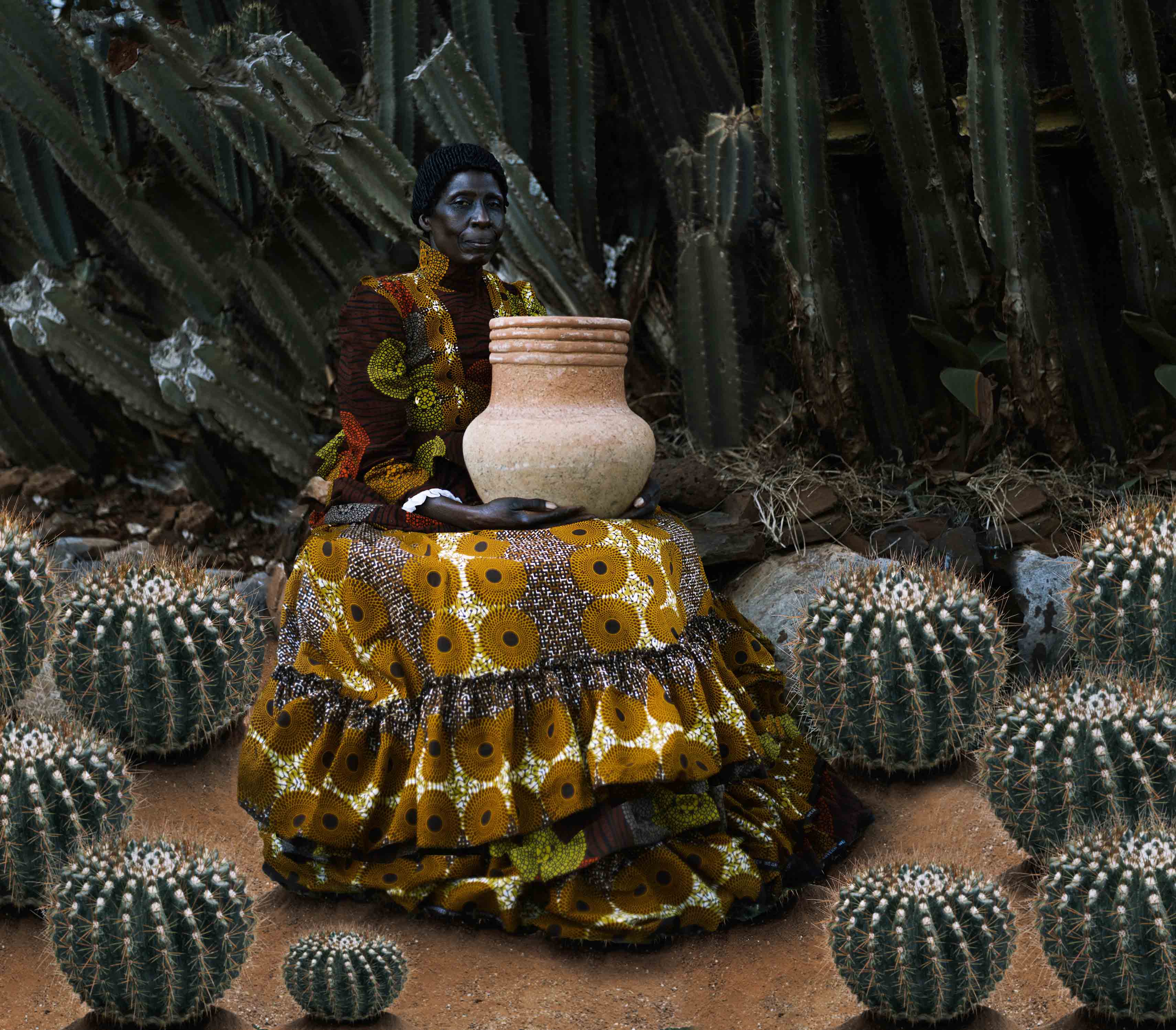A Black woman in a large red and yellow dress holds a large pot among cacti